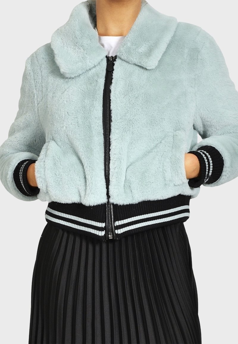 Giacca bomber in pelliccia sintetica azzurro chiaro con chiusura frontale a zip, polsini e orlo a costine neri con dettagli a strisce bianche, abbinata a una gonna nera plissettata.