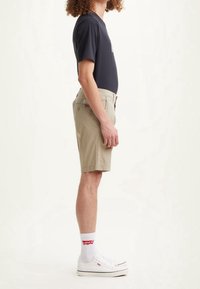 Pantaloni corti beige con una vestibilità rilassata, dotati di tasche laterali e etichetta sul retro. Abbinati a una maglietta scura e scarpe da ginnastica bianche con dettagli rossi.