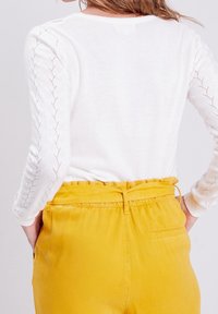 Bílý dlouhý rukáv top s propracovanými pletenými vzory na rukávech, doplněný Vysoce pasovými žlutými kalhotami se zavazováním v pase a postranními kapsami.