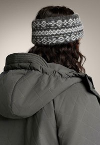 Šedá pletená čelenka s geometrickým vzorom, skombinovaná s tmavozelenou prešívanou bundou. Textúry efektívne kontrastujú.