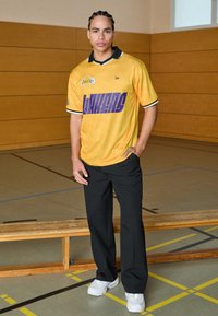 Gelbes Basketballtrikot mit lila "LAKERS"-Schriftzug und schwarzem Kragen, kombiniert mit schwarzer Hose und weißen Turnschuhen, in einer Turnhalle.