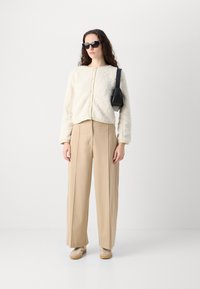 Cream-colored fleece cardigan with button closure, paired with high-waisted beige pleated trousers and light sneakers. Black handbag.