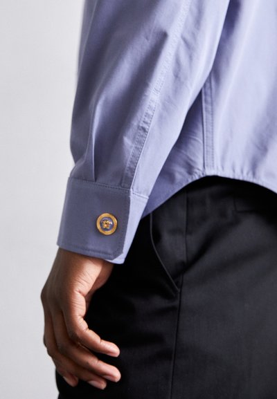 Camisa de manga larga de color púrpura claro con un puño que presenta un botón redondo dorado. Tela suave con un corte ajustado, delimitada por costuras limpias.