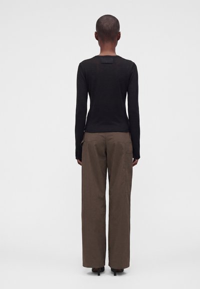 Personne aux cheveux courts portant un haut noir à manches longues, un pantalon large marron et des chaussures à talons noires, debout de dos sur un fond uni.