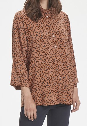 Kvinde iført en brun, langærmet, knappet bluse med sort leopardprint, kombineret med mørke bukser, stående mod en hvid baggrund.