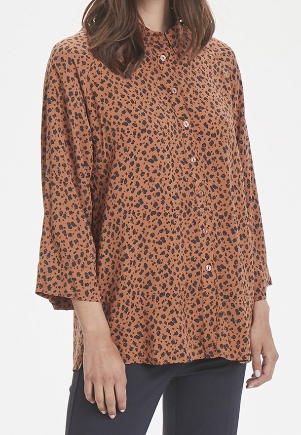Femme portant un chemisier marron à manches longues boutonné avec un imprimé léopard noir, associé à un pantalon foncé, debout devant un fond blanc.