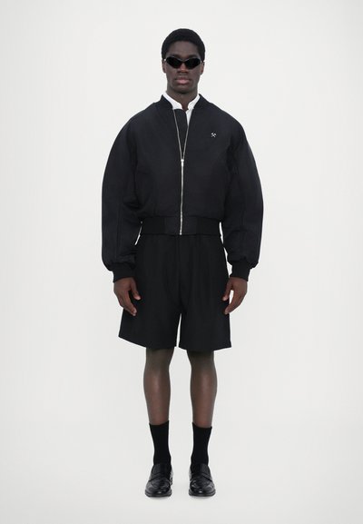Veste bomber noire avec fermeture éclair sur le devant, short noir, chaussettes noires et chaussures noires, avec un design minimal et un tissu texturé.