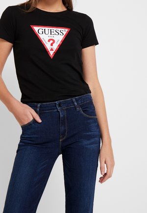 Camiseta negra con un gráfico triangular rojo que presenta "GUESS®", combinada con vaqueros azul oscuro. Mangas cortas, diseño ajustado, estilo casual.