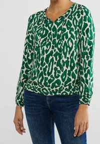 Donna che indossa una blusa a maniche lunghe verde e bianca con stampa leopardata, scollo con cordoncino e jeans blu scuro, in piedi contro uno sfondo neutro.