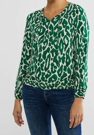 Frau trägt eine grün-weiße Bluse mit Leopardenmuster und langen Ärmeln, mit einer Kordelzug-Ausschnitt, und eine dunkelblaue Jeans, steht vor einem einfarbigen Hintergrund.