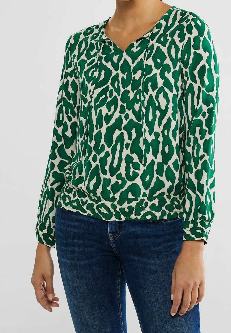 Donna che indossa una blusa a maniche lunghe verde e bianca con stampa leopardata, scollo con cordoncino e jeans blu scuro, in piedi contro uno sfondo neutro.