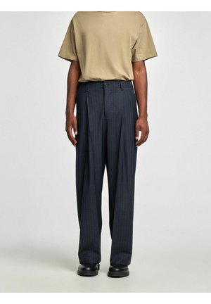 Homme portant un pantalon à rayures fines sombre et ample, une chemise beige à manches courtes et des chaussures noires, debout devant un fond clair uni.