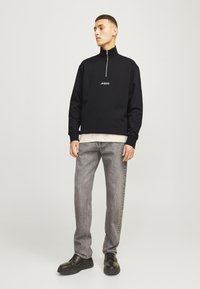 Black zip-up sweatshirt with a high collar and white logo. Paired with light grey jeans and black platform shoes, creating a casual look.