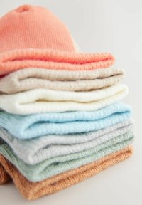 Stak af ni strikkede hatte i pastel farver: koral, beige, creme, lyseblå, grå, mintgrøn og orange, der viser en blød tekstur og enkelt design.