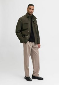 Olivna zelena jakna z več žepi, visokim ovratnikom in zapiranjem na pritisne gumbke, v kombinaciji s smetanastimi hlačami in črnimi čevlji. Gladek, mat videz.