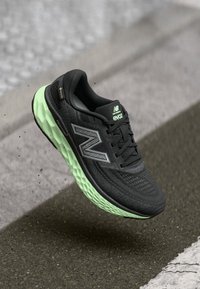 Mustard- ja roheline New Balance jooksutoss õhus asfaltteel, rõhutades hingavat võrkmaterjali ülemust ja pehmendatud taldadesse disaini.