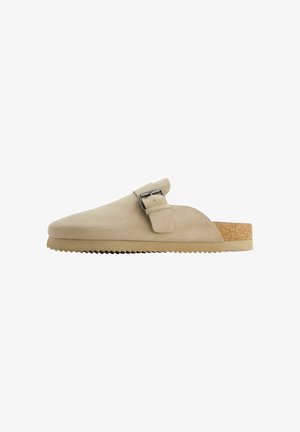 Beige Wildlederschuh mit geschwungener Form, verstellbarem Metallverschlussriemen und strukturiertem Korkfußbett. Die Laufsohle hat eine gezahnte Kante für besseren Halt.