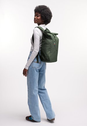 Mujer con cabello rizado, vestida con una camiseta blanca de manga larga y jeans azules, llevando una mochila verde grande, de pie contra un fondo blanco.