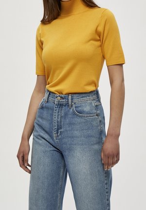 Woman wearing a mustard yellow short-sleeve turtleneck tucked into high-waisted blue denim jeans against a plain background.