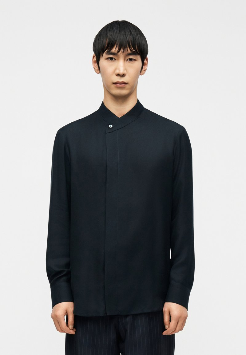 Άνδρας με κοντά μαύρα μαλλιά, φορώντας μαύρο που κάμισο με μανσέτα μανδαρίνου με κουμπιά και παντελόνι με λεπτές ρίγες, στέκεται μπροστά σε ένα απλό φωτεινό φόντο.