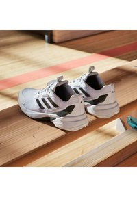 Białe buty sportowe z czarnymi akcentami, z teksturowaną cholewką, wyściółką w okolicy kostki i gumową podeszwą, leżące na drewnianej powierzchni.