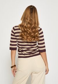 Sweter w paseczki w ciemnobrązowym i beżowym kolorze z rękawami do łokci, zestawiony z beżowymi spodniami. Włosy ułożone w luźne fale.