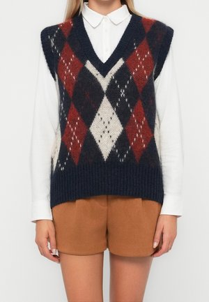 Femme portant un gilet en maille losangé bleu marine, rouge et crème sur une chemise blanche à col avec un short marron clair.