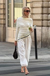 Maglietta bianca a maniche lunghe con design drappeggiato, cintura a pattern attorno alla vita. Pantaloni bianchi larghi, borsa nera, sandali neri, mentre tiene in mano una bevanda verde.
