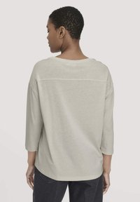 Helle graue Bluse mit dreiviertel Ärmeln. Weicher Stoff und lockere Passform. Verfügt über einen runden Ausschnitt und horizontale Nahtdetails im oberen Rückenbereich.