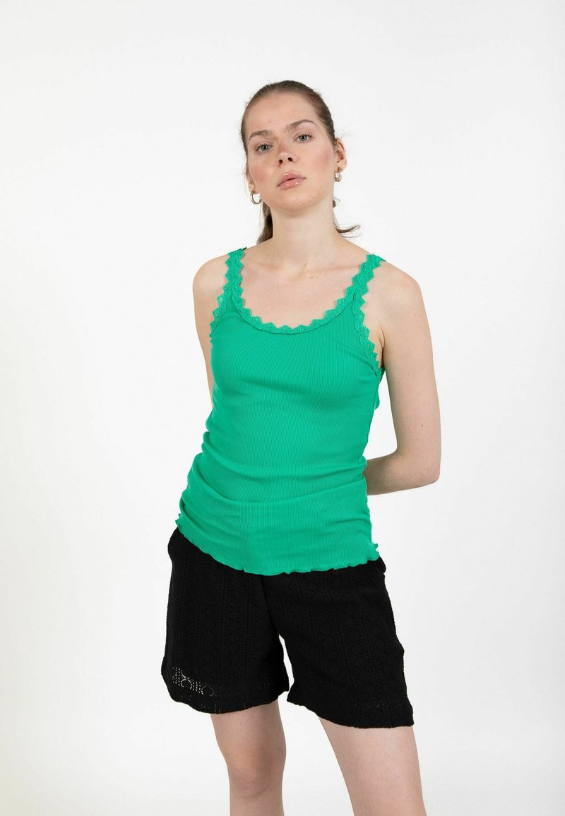 Femme portant un haut vert sans manches aux bords festonnés et un short noir à motif, debout devant un fond blanc uni.