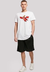 Hvit bomull T-skjorte med et fargerikt krabbe-motiv, kombinert med svarte shorts og hvite joggesko, satt mot en nøytral bakgrunn.
