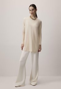 Beige hosszú ujjú felső, kerek nyakkivágással, fehér széles szárú nadrággal párosítva. Arany láncnyaklánc ad egy fémes akcentust. Simább anyagtextúra.