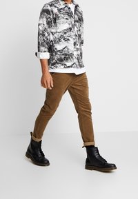 Uomo che cammina indossando un giacca a strisce bianche e nere, una camicia bianca, pantaloni marroni e stivali neri stringati su uno sfondo bianco.