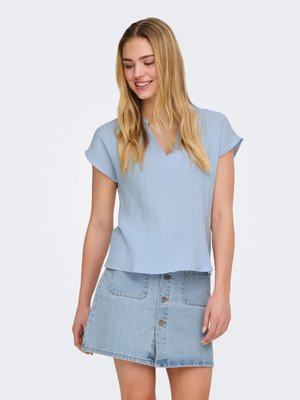 Joven con pelo largo y rubio que lleva una blusa de manga corta azul claro y una falda vaquera con botones al frente, sonriendo y mirando hacia abajo.