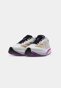 Scarpe da corsa con tomaia in rete bianca, strisce dorate, dettagli neri e accenti viola. Presentano suole spesse e imbottite con battistrada testurizzato.