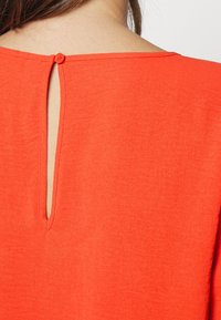 Haut rouge à manches courtes avec une ouverture en forme de trou à l'arrière fermée par un petit bouton, fabriqué dans un tissu léger et lisse avec une texture subtile.