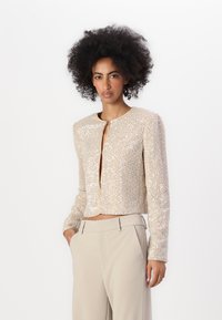 Giacca cropped beige con maniche lunghe, coperta di paillettes. Caratterizzata da un collo rotondo e una piccola chiusura frontale. Indossata con pantaloni beige.