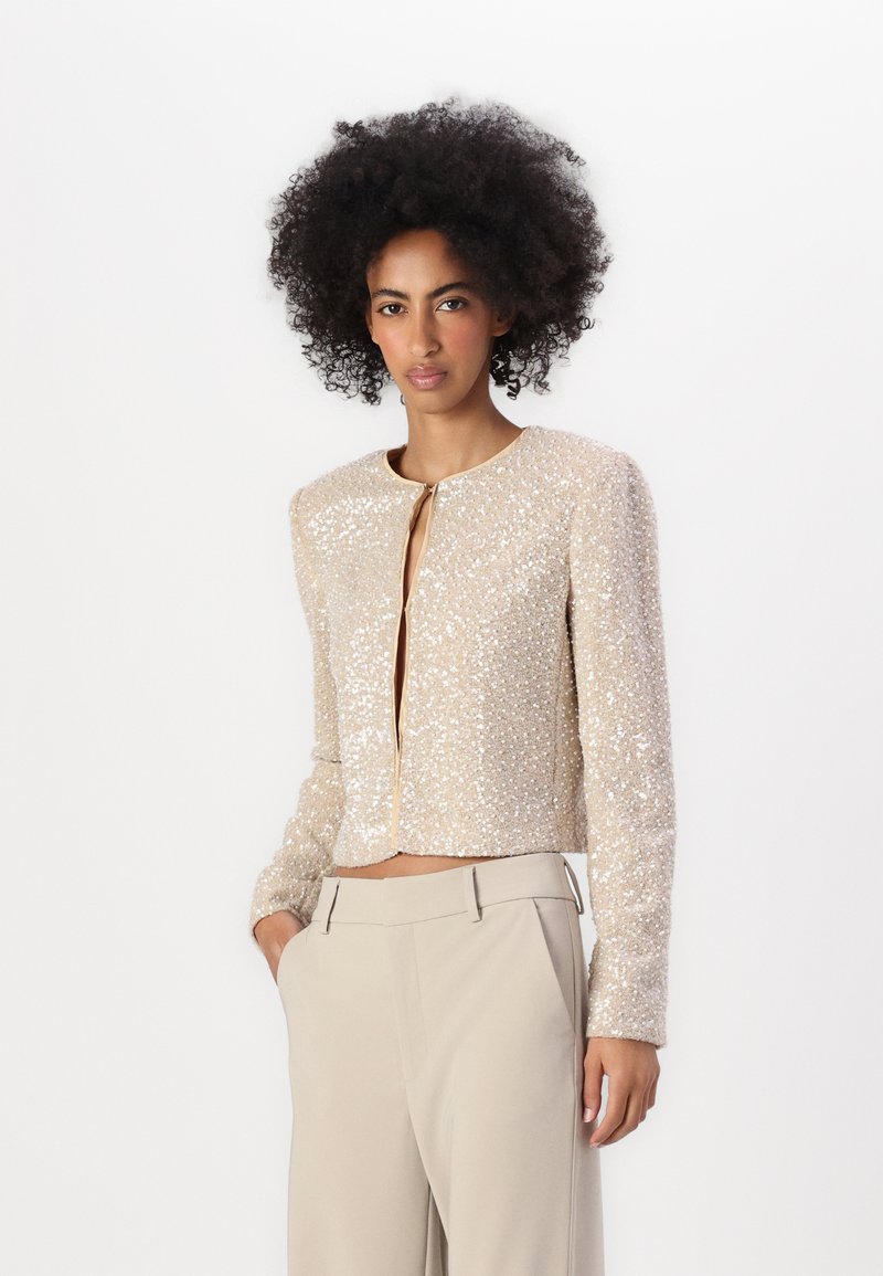 Giacca cropped beige con maniche lunghe, coperta di paillettes. Caratterizzata da un collo rotondo e una piccola chiusura frontale. Indossata con pantaloni beige.