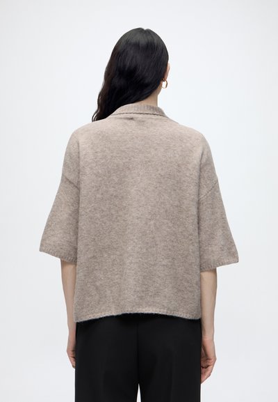 Femme aux longs cheveux foncés portant un pull beige à manches courtes et un pantalon noir, vue de dos sur un fond clair uni.