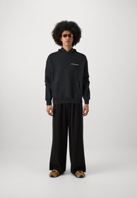 Schwarzer Kapuzenpullover mit einer Vordertasche und weißem Text, kombiniert mit lockeren schwarzen Hosen und gemusterten Schlappen. Braune Sonnenbrillen getragen.