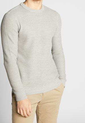 Homme portant un pull en maille texturée gris clair et un pantalon beige, debout avec la main gauche dans la poche, devant un fond uni.