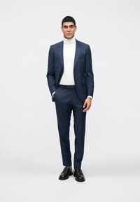 Námornícky oblek s priliehajúcim strihom, skombinovaný s bielym rolákom. Hladká tkanina, úzke nohavice a čierne topánky dokončujú vzhľad. Minimalistický štýl.