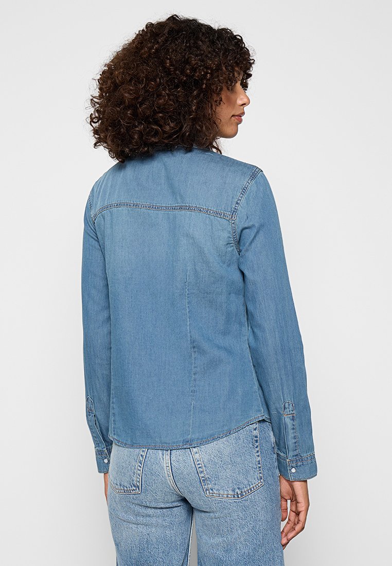 Veste en jean bleu clair avec un col pointu, des manches longues et un ourlet incurvé. Présente un design cousu le long des épaules.
