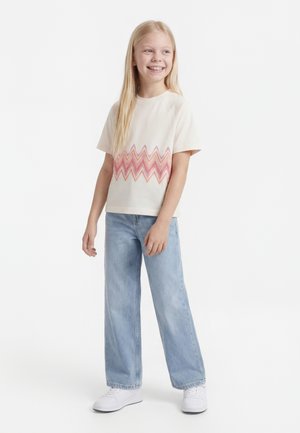 Smilende jente med langt blond hår iført en kremfarget skjorte med rosa sikksakkmønster, lyseblå vide jeans og hvite joggesko.