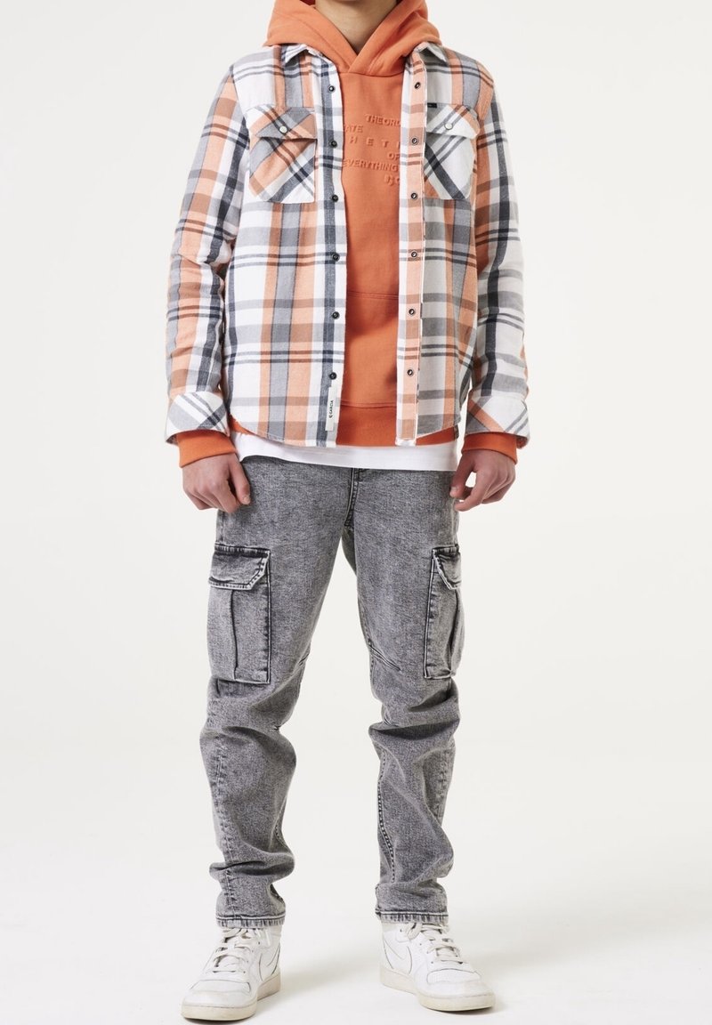 Persona che indossa un hoodie arancione, una camicia a quadri arancione e bianca con bottoni, jeans cargo grigi e sneakers bianche, in piedi contro uno sfondo bianco.