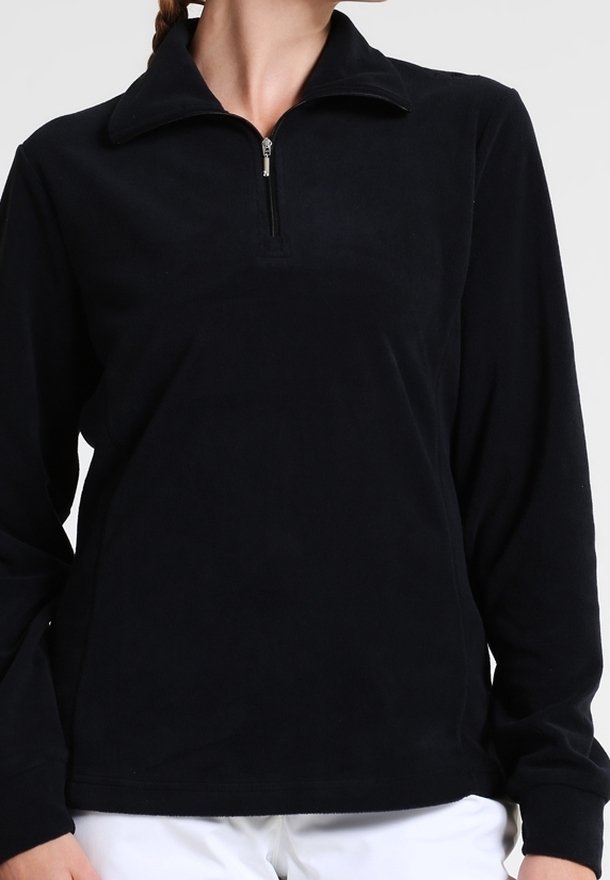 Femme portant un pull noir à manches longues avec une fermeture éclair avant sur un quart et un col, associé à un pantalon blanc.