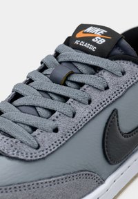Grå Nike SB Classic-sneakers har teksturert skinn og semsket skinn, med grå snører og en svart tunge merket med Nike-logoen og oransje detaljer.
