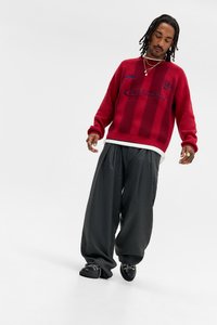 Camisola vermelha de malha com riscas horizontais, com um logótipo. Usada com calças cinzentas largas e sapatos pretos, criando um aspeto em camadas.