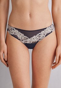 Marinblå bikiniunderdelar med spetsdetaljer i ett blommönster längs midjan och genomskinliga mesh-sidor. Mjuk tyg med en slät yta.