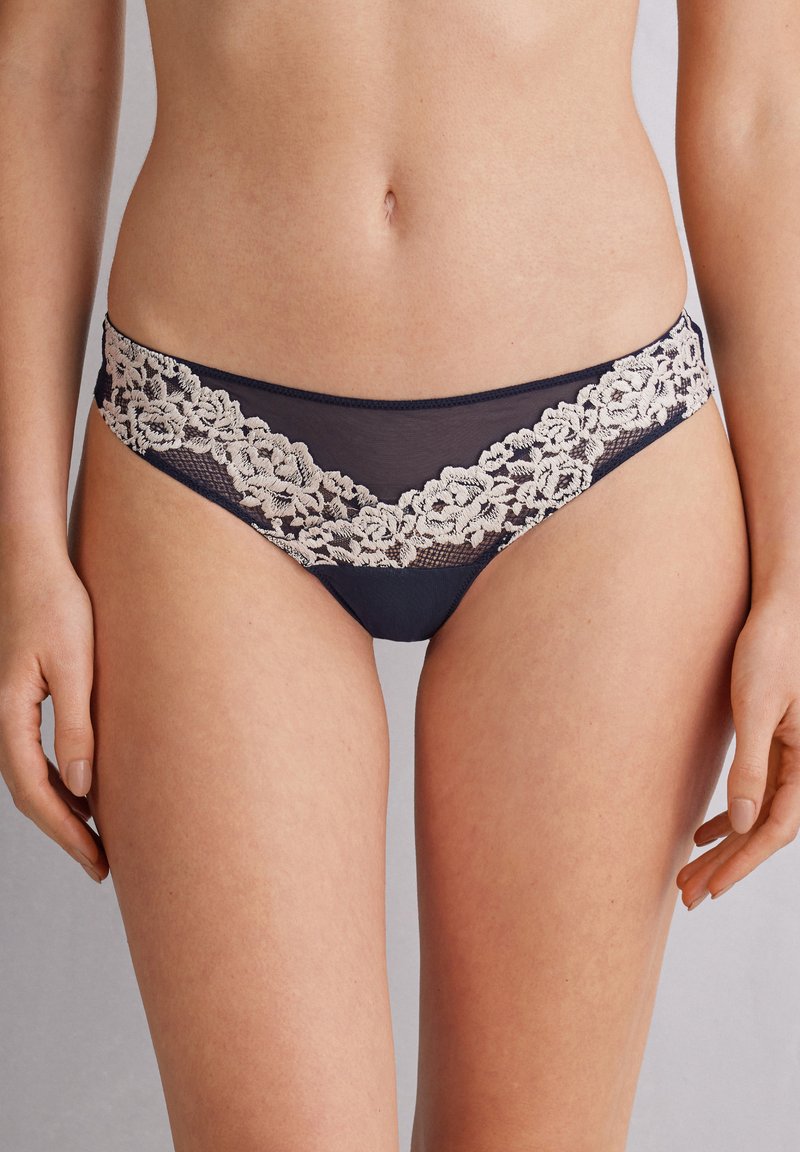 Marinblå bikiniunderdelar med spetsdetaljer i ett blommönster längs midjan och genomskinliga mesh-sidor. Mjuk tyg med en slät yta.